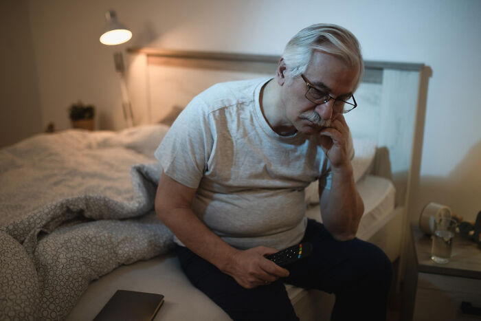 An older man with glasses sits on a bed in a dimly lit room, holding a remote, reflecting on unsettling childhood memories.