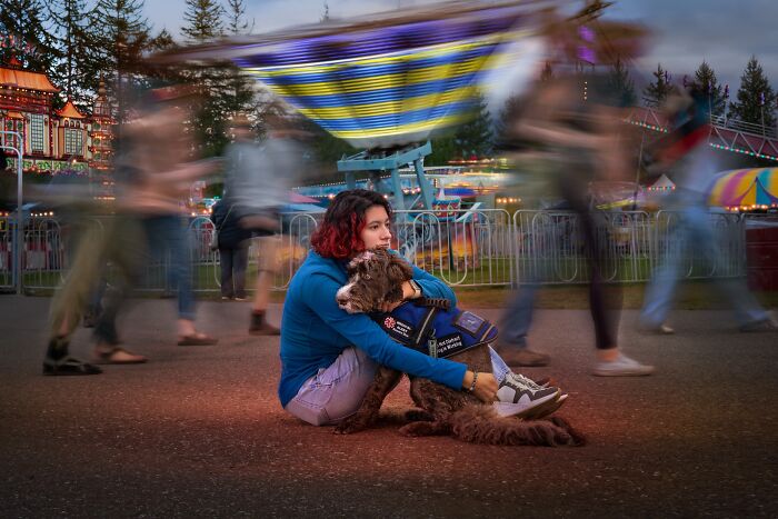 A girl with red hair sits on the ground hugging a brown dog wearing a vest, at a carnival, for pet photography awards.