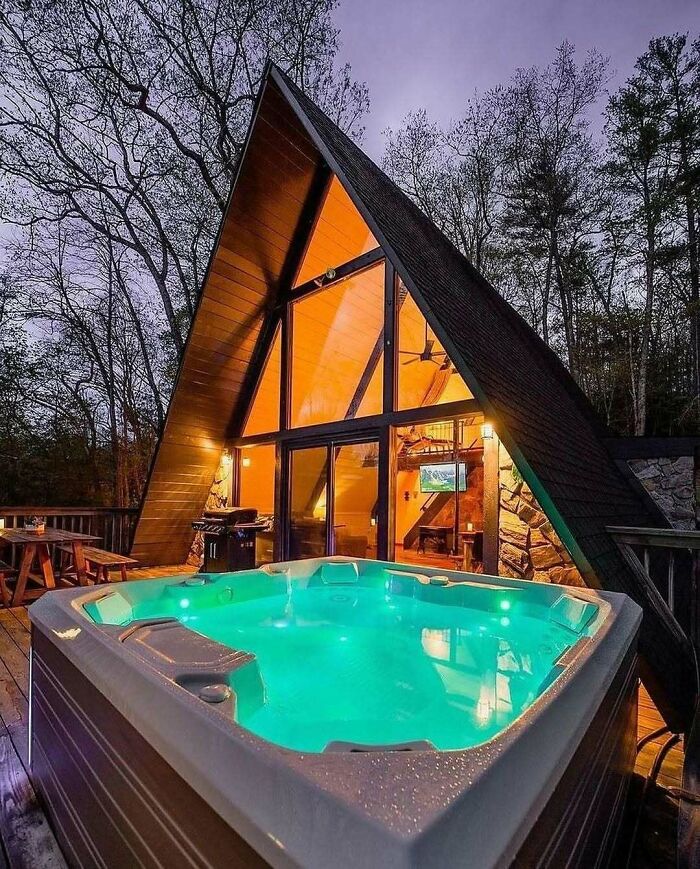 An A-frame house with a brilliant design and large windows, a hot tub on the deck at dusk.