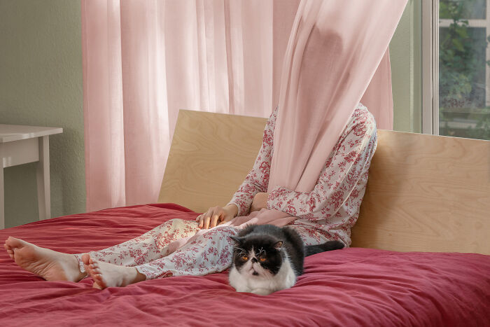 A photographer creates surreal images of a person under a curtain with a cat on a bed.