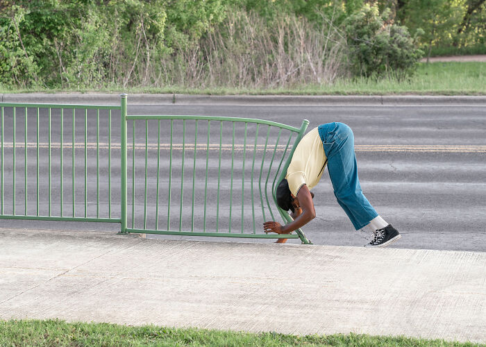 A photographer creates surreal images; a person bends through a green fence on a sidewalk, creating an uncanny scene.