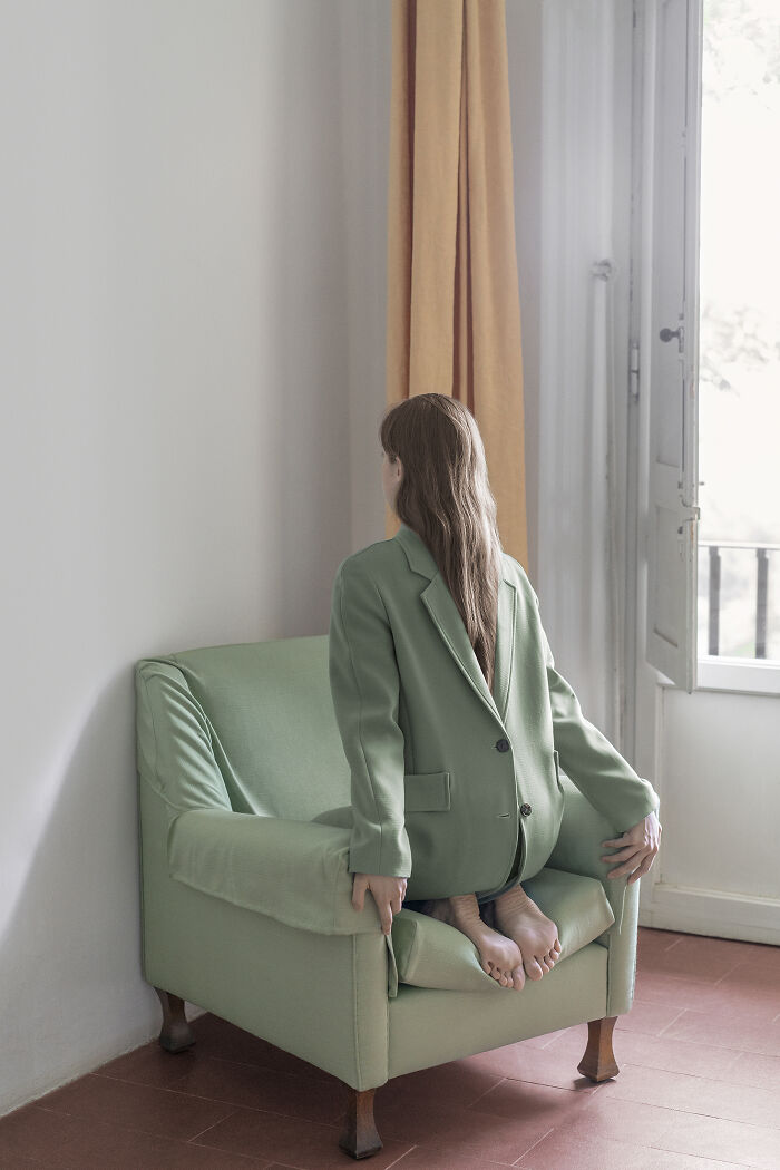 Surreal images: A woman in a green suit kneels on a matching armchair, looking out an open door, making the everyday feel uncanny.