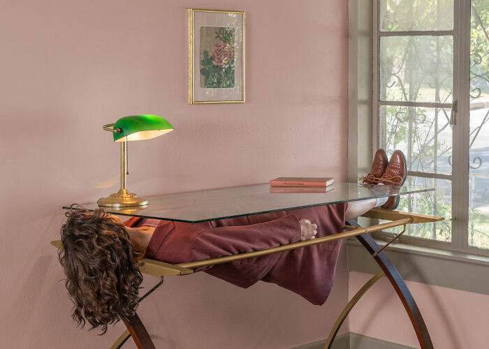 A photographer creates surreal images; a person lies face down on a glass desk in an uncanny, pink room with a green lamp.
