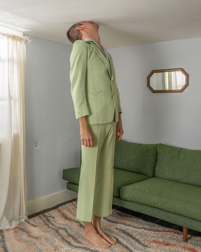 A man in a green suit with his head pressed against the ceiling, presenting a surreal image in an everyday room.