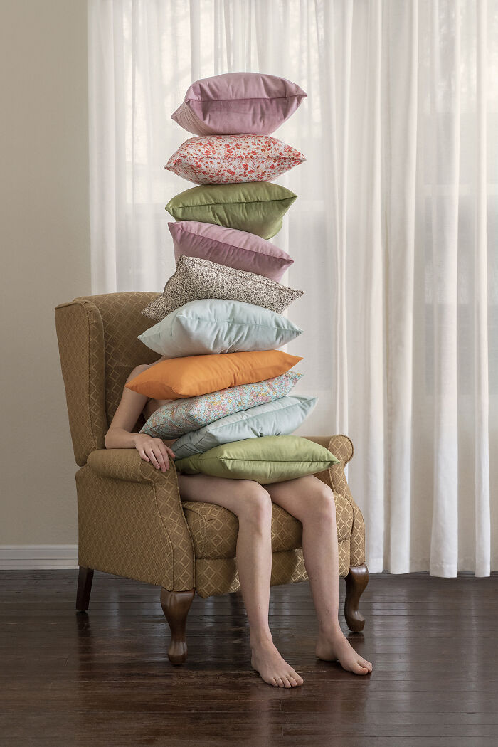 A person hidden behind a tall stack of pillows on an armchair, creating a surreal image.