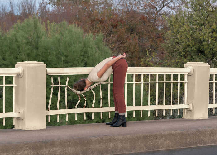 A woman in brown pants and a light shirt bends deeply from the waist over a bridge railing in a surreal image.