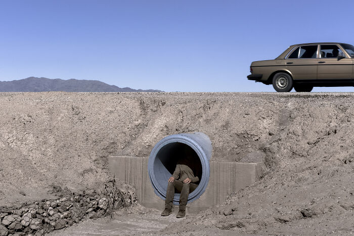A person is sitting inside a large pipe by the road as a car drives by, creating an uncanny, surreal image.