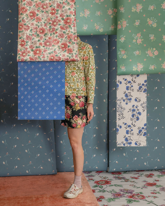 A woman stands amidst floral wallpaper, blending in with her patterned clothing, creating a surreal image.