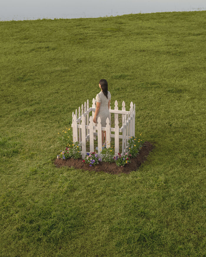 A photographer creates surreal images of a woman standing in a small white picket fence in a grassy field.