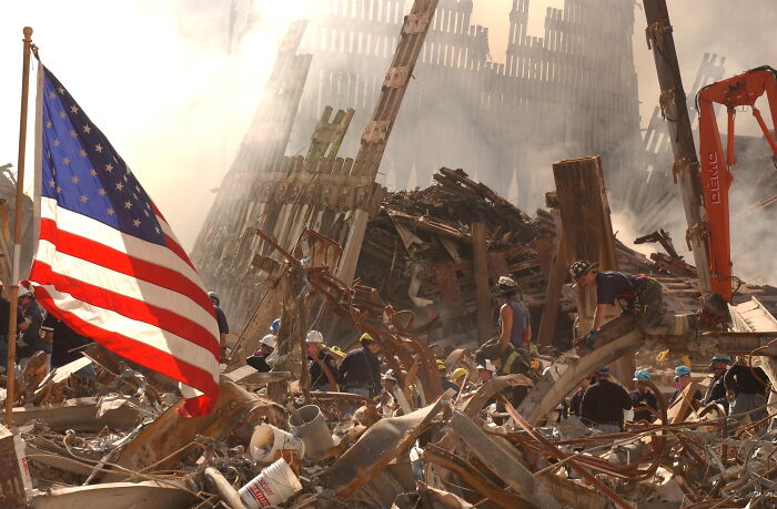 An American flag stands proudly amidst the 9/11 wreckage of the Twin Towers, with rescue workers searching for survivors.