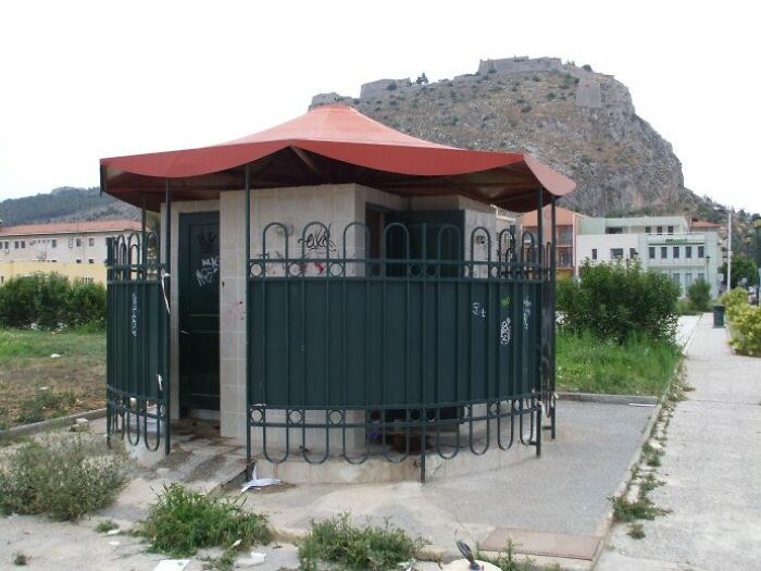 Urban designers forgot about people's comfort; a public restroom with an oddly shaped roof and fencing, under a distant castle.