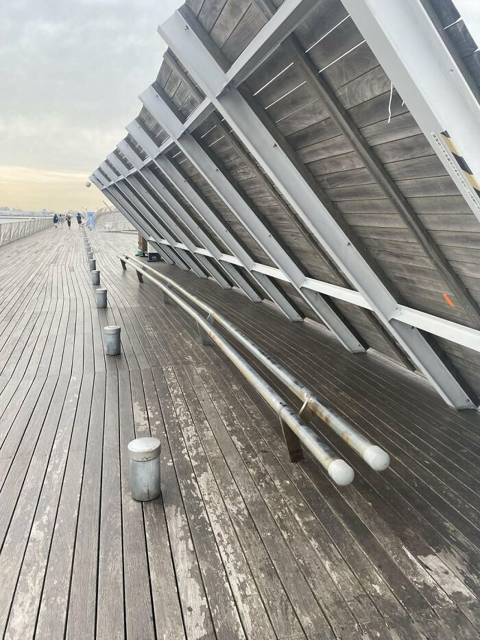 A long wooden walkway with a slanted metal structure overhead, featuring a bench made of two metal pipes, highlighting urban design issues.
