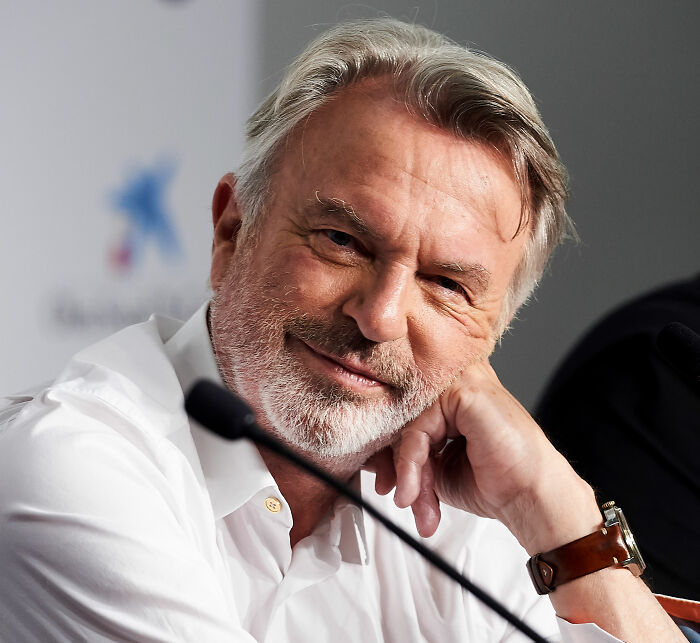 A smiling Sam Neill wearing a white shirt, resting his head on his hand. Fans debate Alec Baldwin tribute. A smiling Sam Neill wearing a white shirt, resting his head on his hand. Fans debate Alec Baldwin tribute.