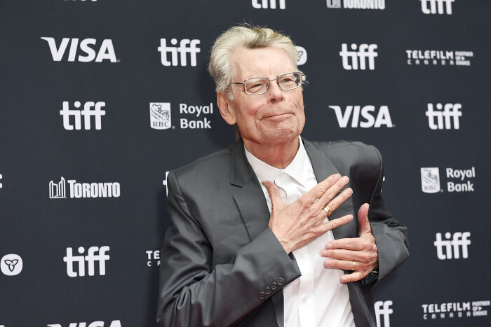 Stephen King, an older man with glasses, in a gray suit, posing on a red carpet. Stephen King, an older man with glasses, in a gray suit, posing on a red carpet.