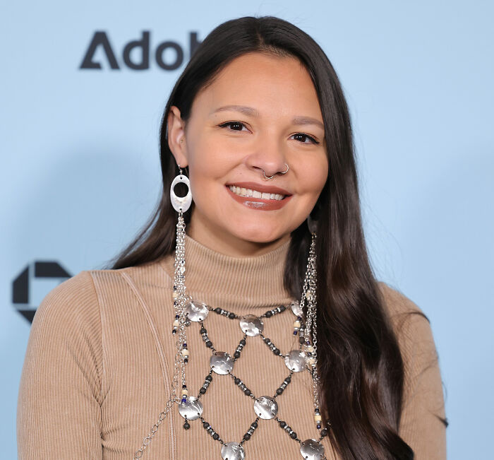 A smiling woman with long dark hair, wearing a beige turtleneck and elaborate silver jewelry, looking at the camera. She is the girlfriend of the alleged cheater.