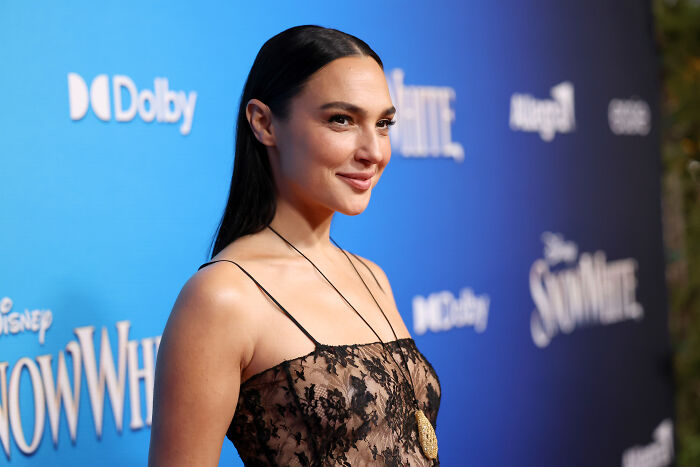 Gal Gadot at a red carpet event, smiling softly, with a dark blue background and event branding.