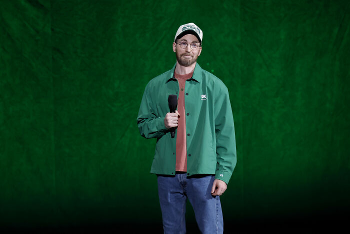 Chris Evans with a glowdown, wearing a green jacket, glasses, and a hat, holding a microphone. Chris Evans with a glowdown, wearing a green jacket, glasses, and a hat, holding a microphone.