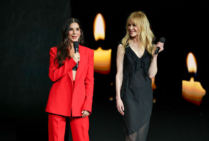 Original actress in red suit speaks with a microphone, next to another actress in a black dress, both smiling, '90s cult classic sequel. Original actress in red suit speaks with a microphone, next to another actress in a black dress, both smiling, '90s cult classic sequel.