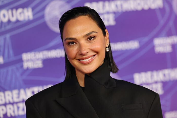 Gal Gadot smiles, wearing a black blazer, against a purple background, her career and new Netflix film in focus.