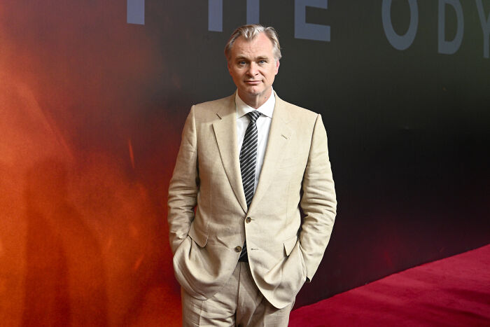 Christopher Nolan, director, in a light-colored suit and striped tie, stands formally in front of an orange-red background. Christopher Nolan, director, in a light-colored suit and striped tie, stands formally in front of an orange-red background.