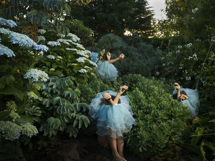 Multiple figures in light blue dresses are interspersed among dense green bushes and flowers, making surreal photos.