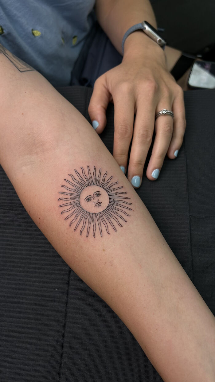 A professional tattoo artist's work: a small, detailed sun tattoo on a woman's forearm.