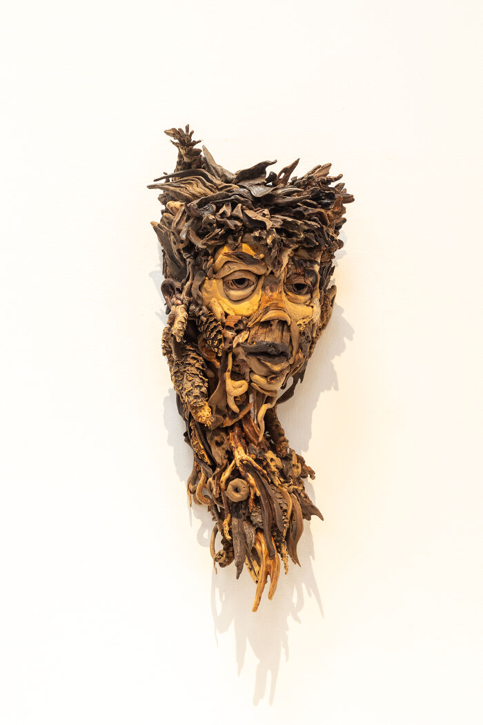A detailed wood sculpture of a somber face and neck, created from collected pieces of wood, mounted on a white wall.