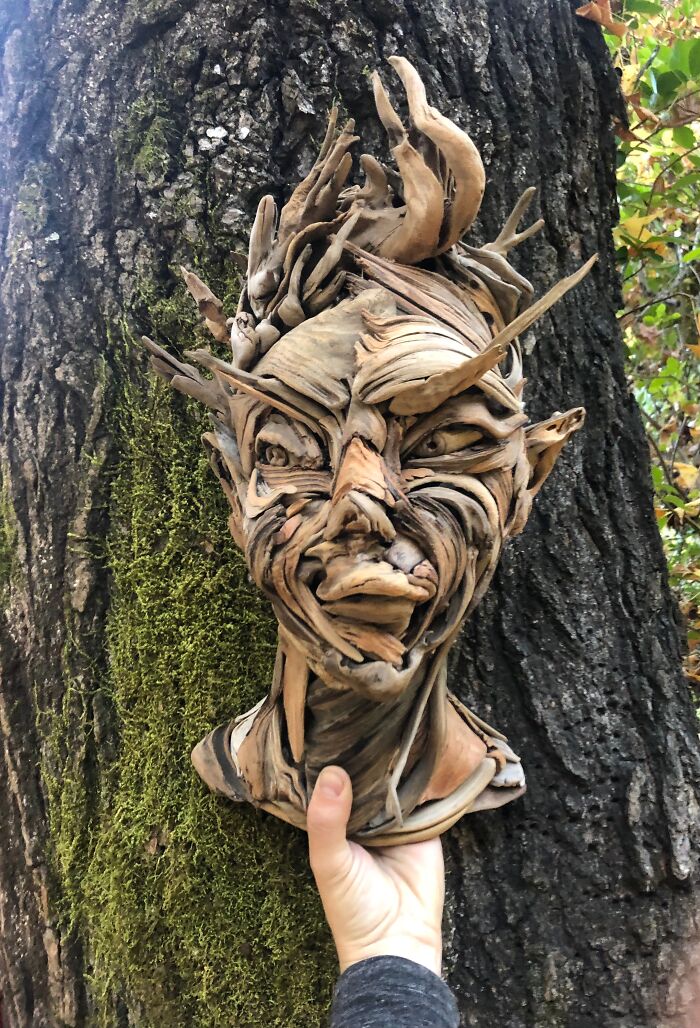A hand holding up a striking wood sculpture of a face with elven features, made from pieces of wood, against a tree.