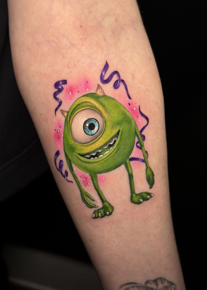 Mike Wazowski tattoo with purple streamers on an arm. Professional tattoo artist renders animated characters.