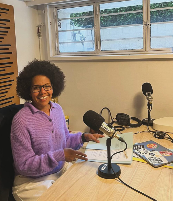 Paris Deputy Mayor, a woman with an afro, smiles while sitting at a radio studio desk, discussing Europe's climate crisis. Paris Deputy Mayor, a woman with an afro, smiles while sitting at a radio studio desk, discussing Europe's climate crisis.