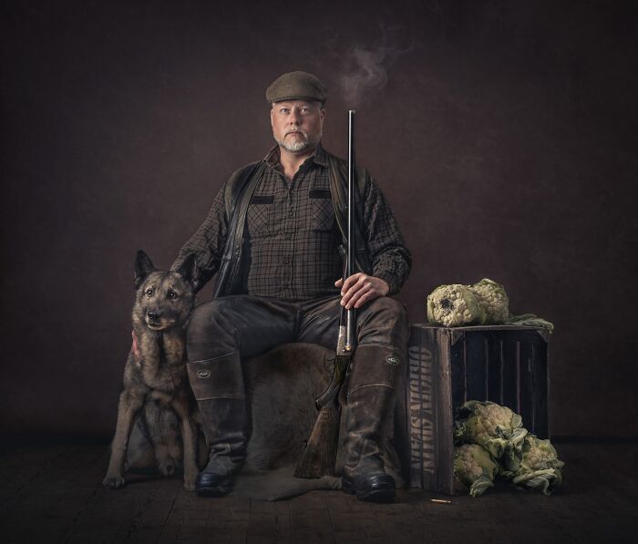 A hunter with a rifle sits next to his dog, with vegetables and a wooden crate, for International Pet Photography Awards.