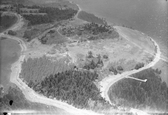 An aerial black and white photo of a landmass with trees and roads next to water. It hints at unsolved mysteries.