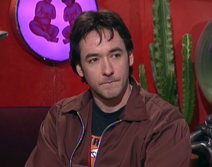 John Cusack in a brown jacket, appearing thoughtful about his vanishing from Hollywood and sister Joan's return.