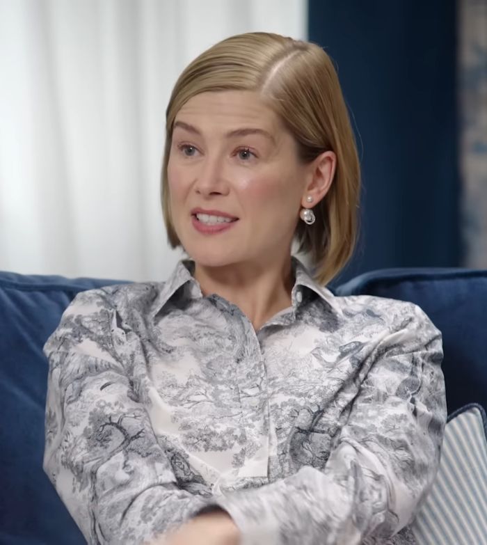 Rosamund Pike, with short blonde hair and pearl earrings, smiles while sitting on a blue couch.