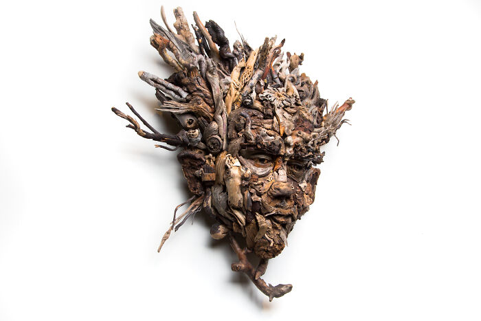 Intricate wood sculpture of a face and head, crafted from collected natural wood pieces, with a dark eye and textured surface.