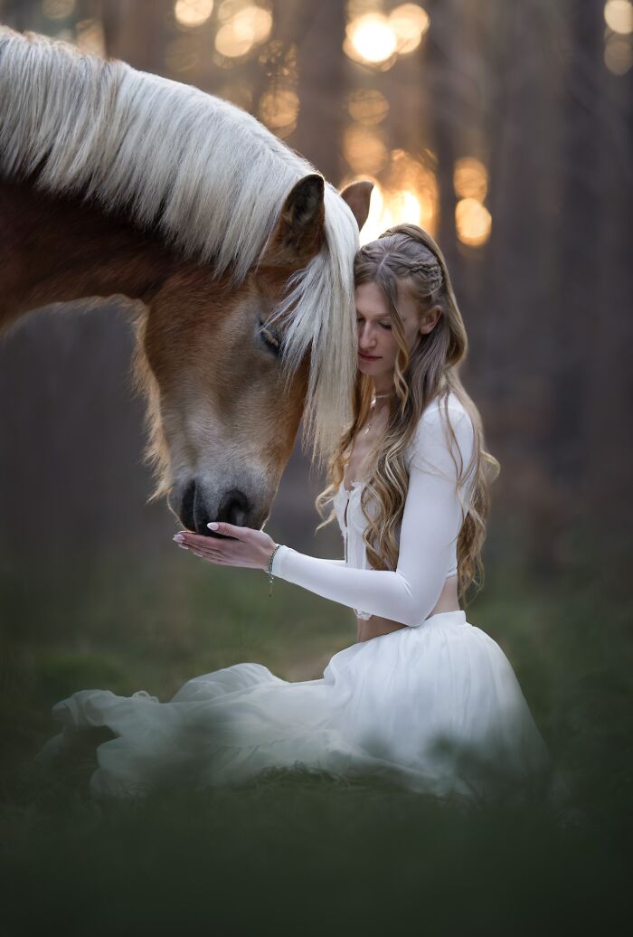 A woman in white dress kneeling, gently touching a horse's head in a forest, for International Pet Photography Awards.