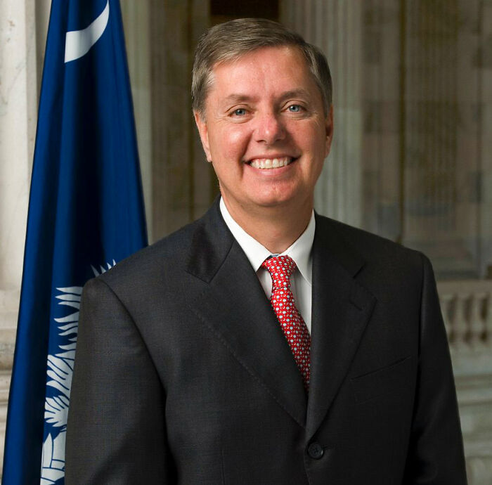 Senator Lindsey Graham smiling against a blue flag, facing allegations from a trans influencer.
