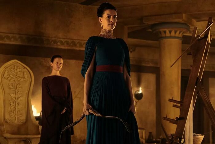 A woman in a teal dress holding a curved object, with another woman behind her. Christopher Nolan's bizarre rules on set. A woman in a teal dress holding a curved object, with another woman behind her. Christopher Nolan's bizarre rules on set.