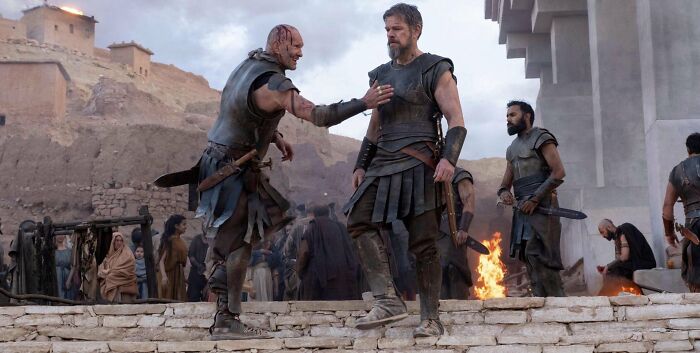 Two armored men on a stone platform, one reaching out, amid a crowd and burning structures in The Odyssey. Two armored men on a stone platform, one reaching out, amid a crowd and burning structures in The Odyssey.