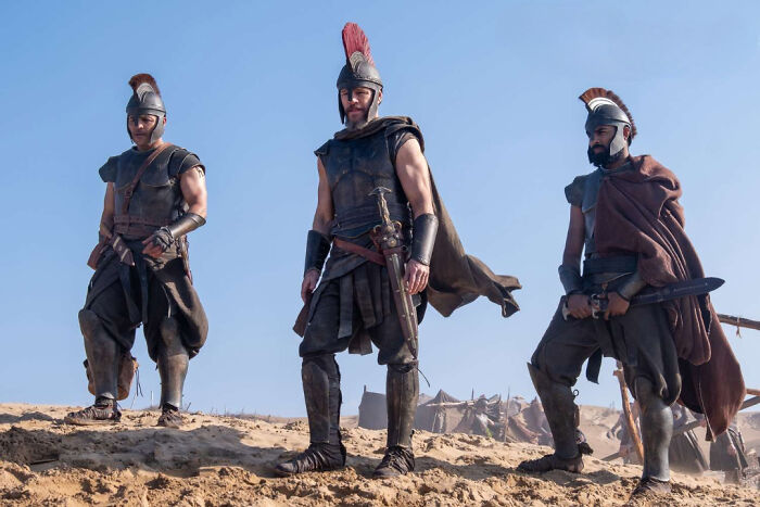 Three warriors in ancient Greek attire stand on a sandy terrain from The Odyssey. Three warriors in ancient Greek attire stand on a sandy terrain from The Odyssey.