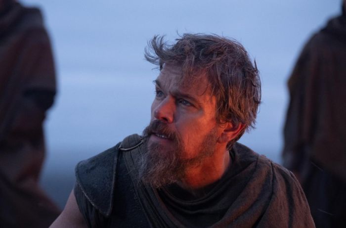 A bearded man, looking intently upward, from The Odyssey adaption, amidst a dramatic, hazy backdrop.