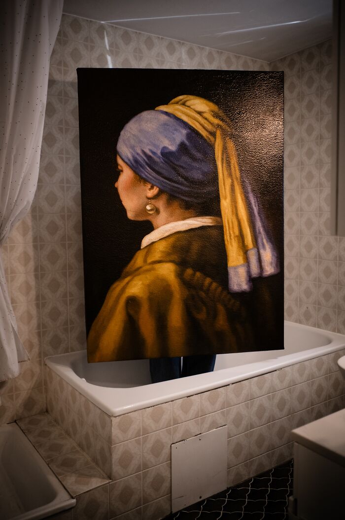 A person holds a reimagined masterpiece painting of Girl with a Pearl Earring from behind, standing in a bathtub.
