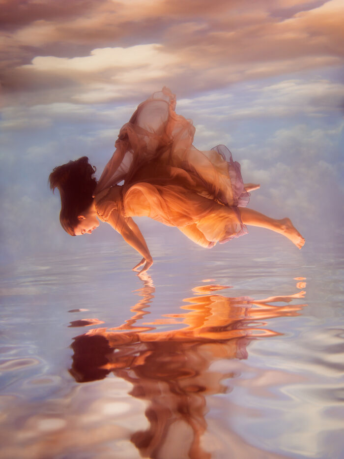 A woman in a flowing dress creates surreal photos by submerging herself in water, gracefully touching her reflection.