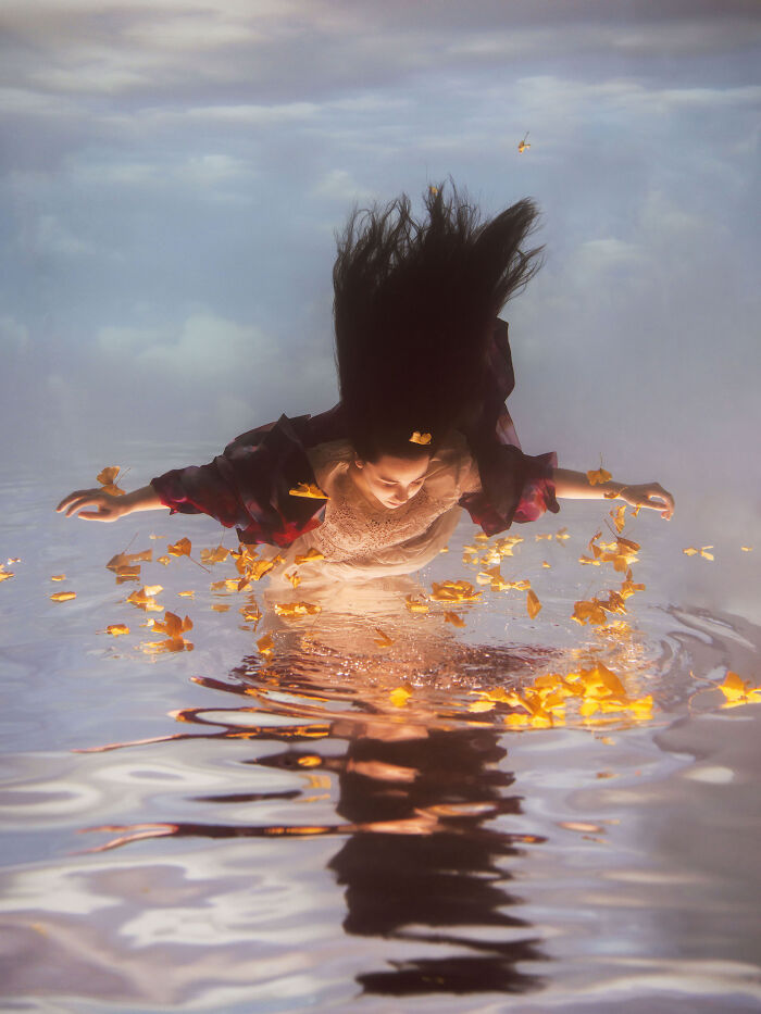 A woman with long dark hair creates surreal photos, submerged in water with autumn leaves and her reflection.