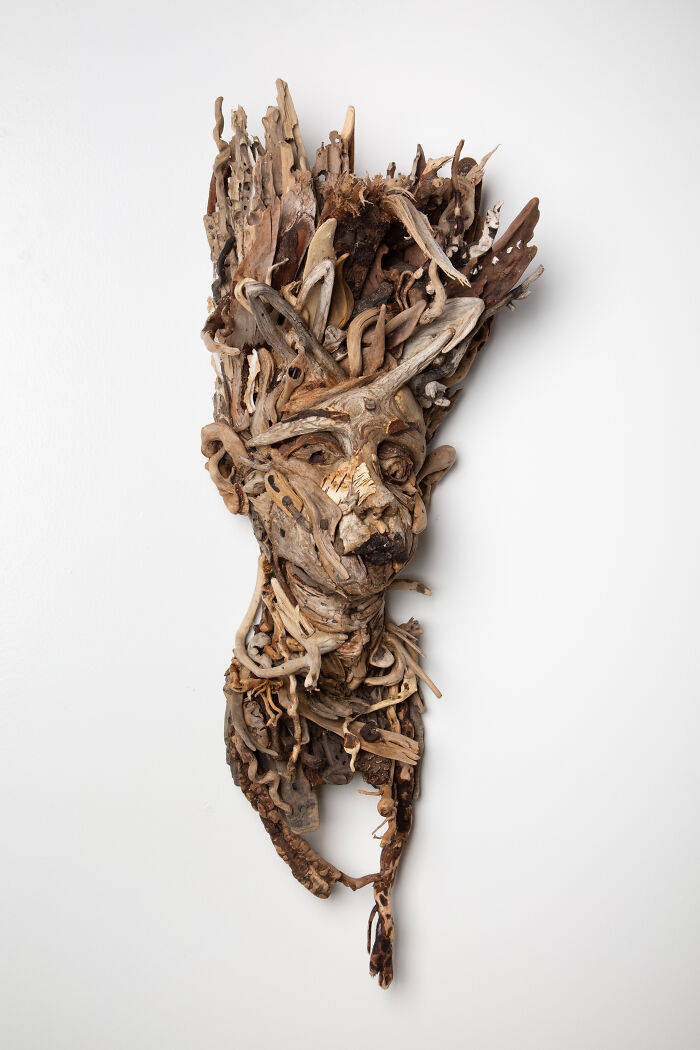 Elongated wood sculpture of a face and upper torso made from collected natural wood pieces, against a white background.