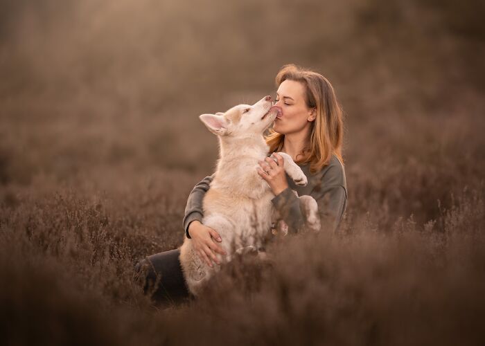 A woman sits in a field of heather, affectionately kissing a light-colored dog, from International Pet Photography Awards.