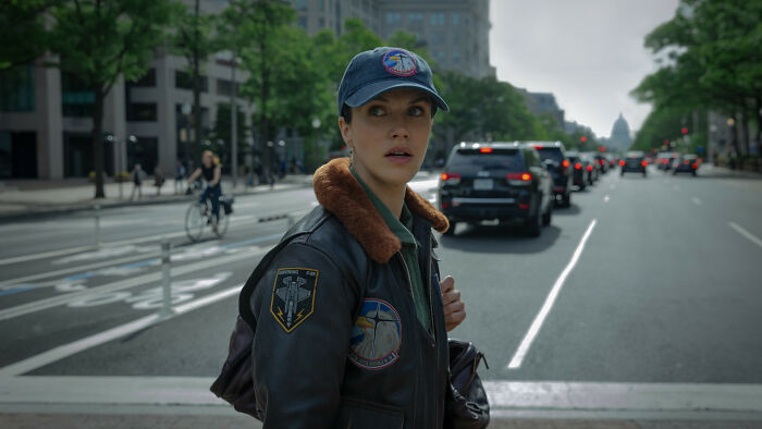 A female Silo Season 3 cast member in a pilot's jacket and cap looks surprised, standing on a city street. A female Silo Season 3 cast member in a pilot's jacket and cap looks surprised, standing on a city street.