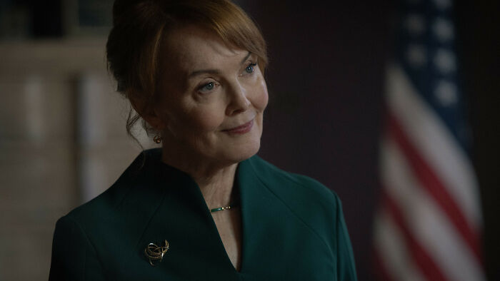 A smiling female Silo Season 3 cast member in a green dress with an American flag in the background. A smiling female Silo Season 3 cast member in a green dress with an American flag in the background.