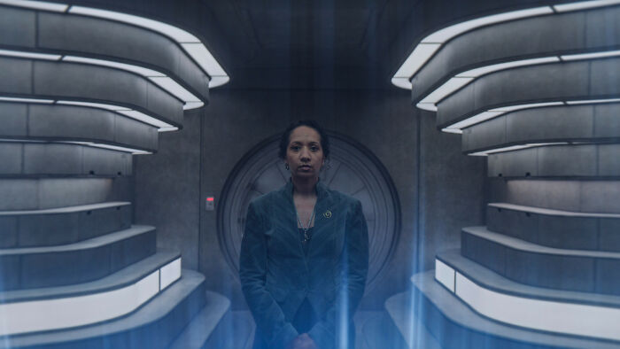 A woman with dark hair, a returning character in the Silo Season 3 cast, stands in a futuristic, sterile chamber. A woman with dark hair, a returning character in the Silo Season 3 cast, stands in a futuristic, sterile chamber.