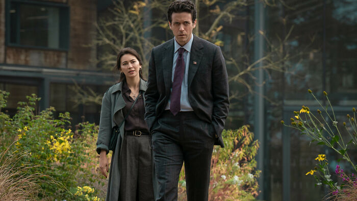 Two Silo Season 3 cast members walk side-by-side, a man in a suit and a woman in a long coat, amidst greenery. Two Silo Season 3 cast members walk side-by-side, a man in a suit and a woman in a long coat, amidst greenery.